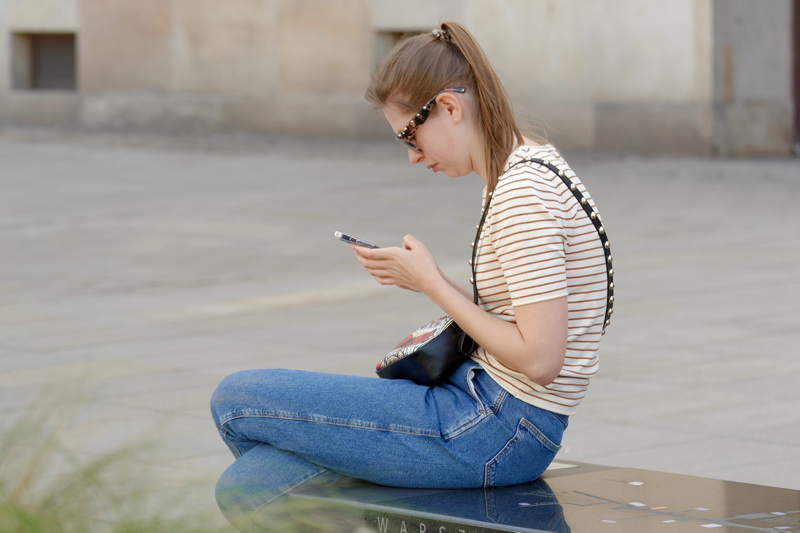 29 - Young woman sitting on a public bench in a city setting while looking down at a smartphone